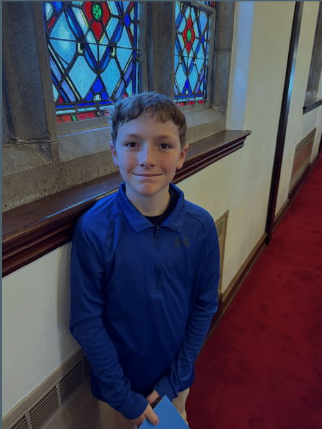 Smiling boy in a blue long-sleeve shirt stands in a hallway with a decorative stained-glass window behind him and a red carpeted floor.