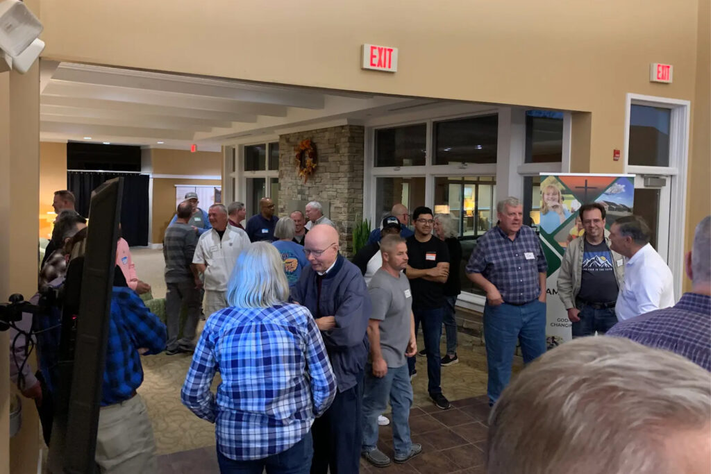 Group of adults chatting and mingling in a spacious indoor lobby at an event, with an exit sign and stone accent wall visible behind them.