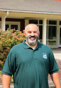Smiling man with a gray beard wearing a dark green polo shirt stands outside a building with pink flowering bushes in the background.