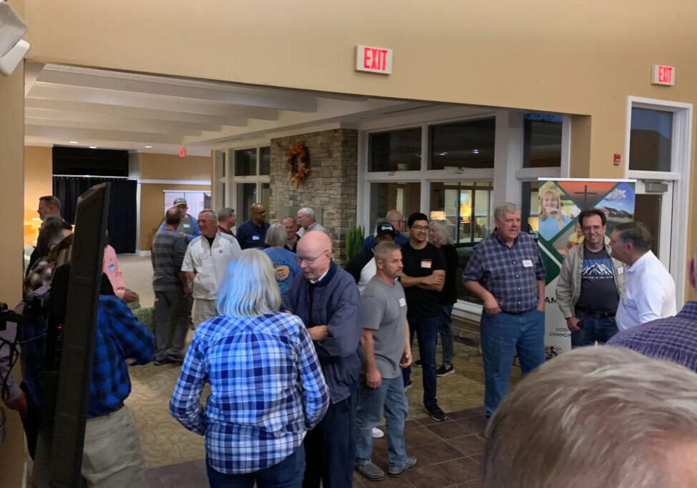 Group of adults chatting and mingling in a spacious indoor lobby at an event, with an exit sign and stone accent wall visible behind them.